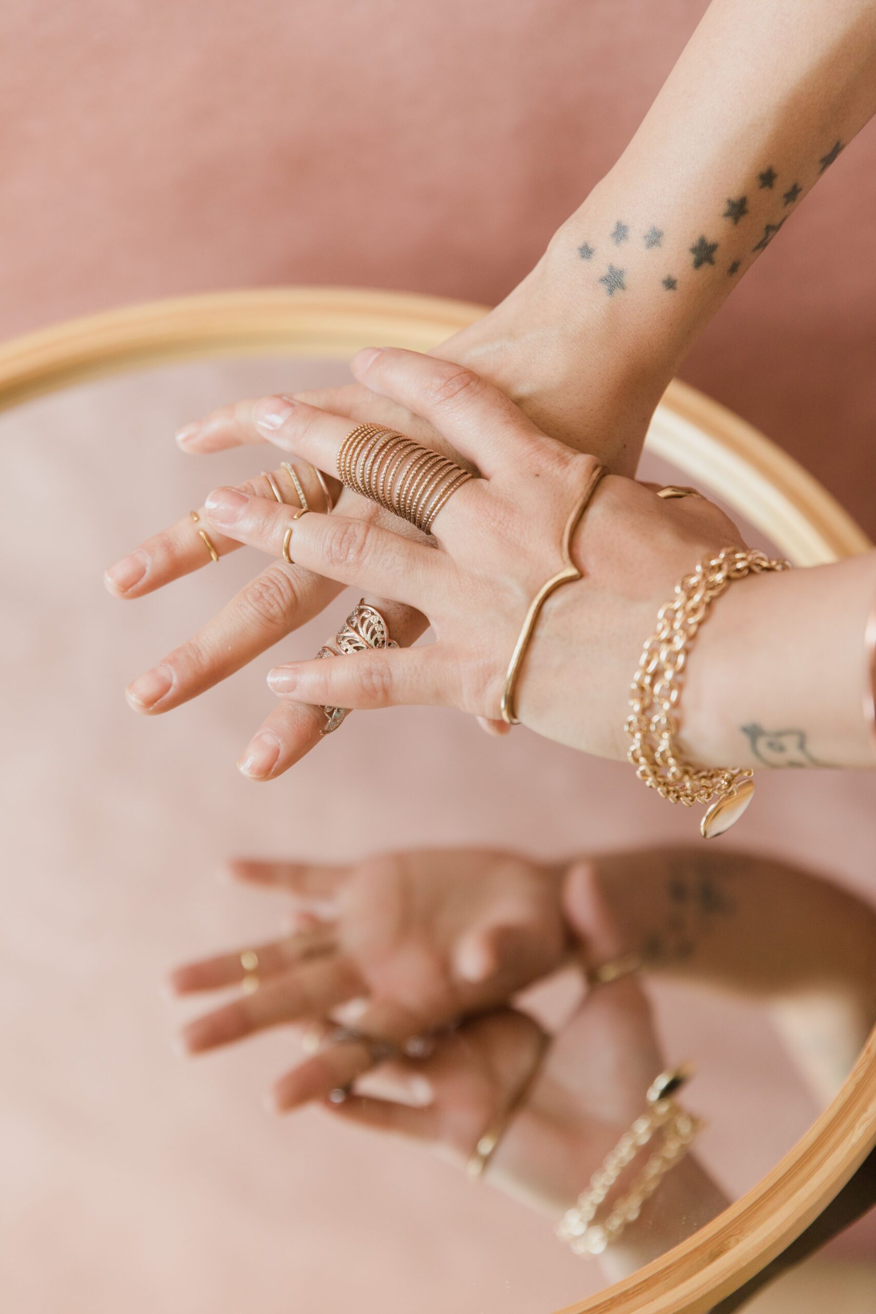 Close-up of tattooed hands with gold rings and bracelets on a reflective surface.