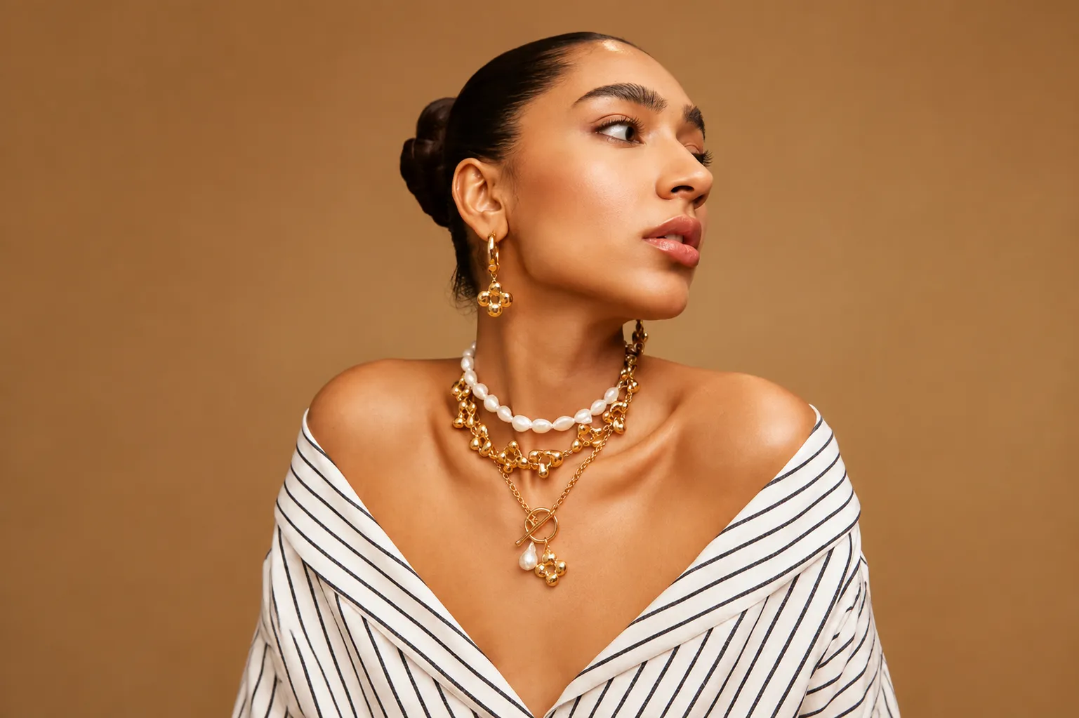 Home A model posing in a striped off-the-shoulder shirt wearing layered gold and pearl jewelry alongside a silver necklace women.