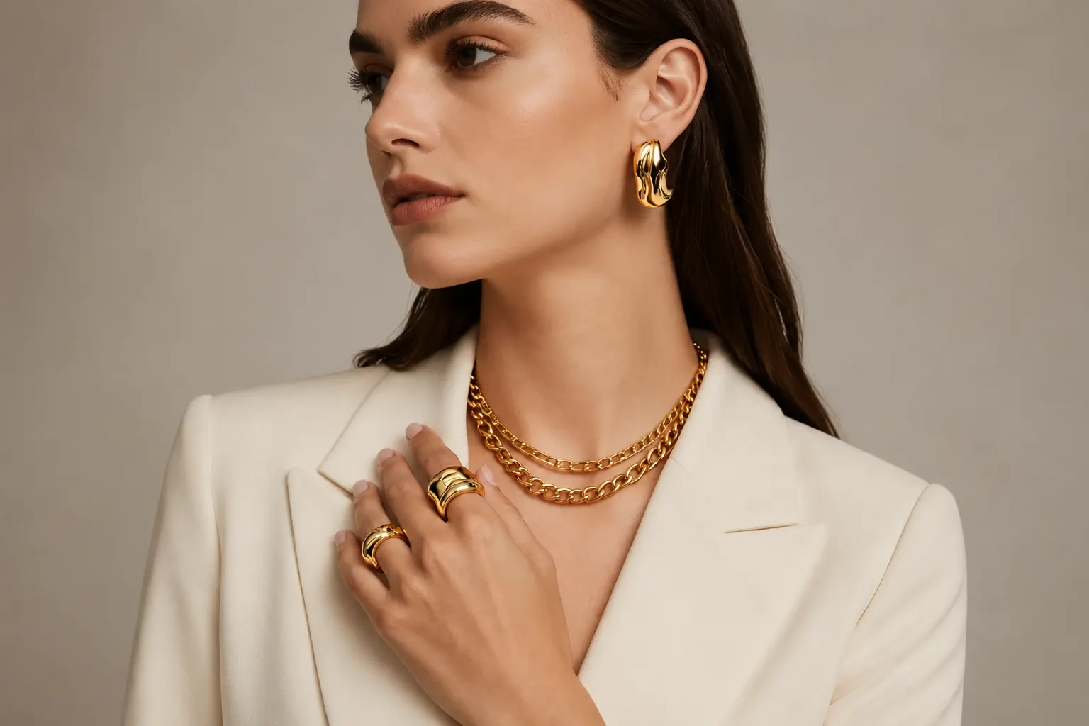 Home A model in a tailored cream blazer wearing bold gold chunky rings, sculptured hoop earrings, and a layered gold necklace.